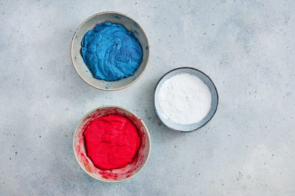 Three bowls on a light surface: one with blue mixture, one with red mixture, and one with white powder.