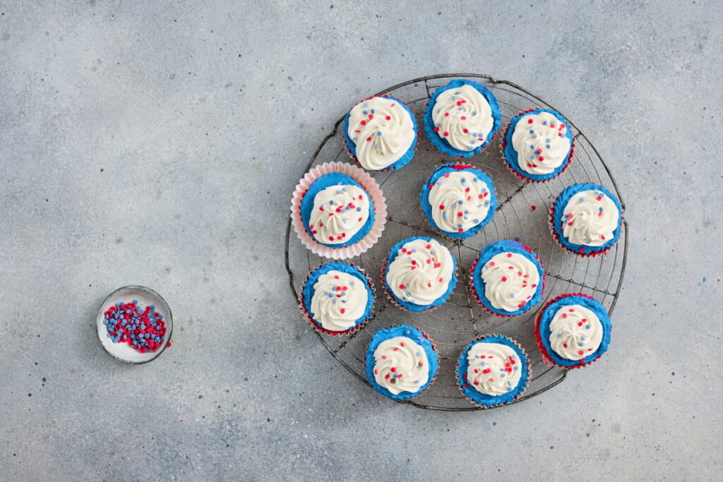 Twelve cupcakes with white frosting and red and blue sprinkles on a cooling rack, with extra sprinkles in a small bowl on a gray surface.