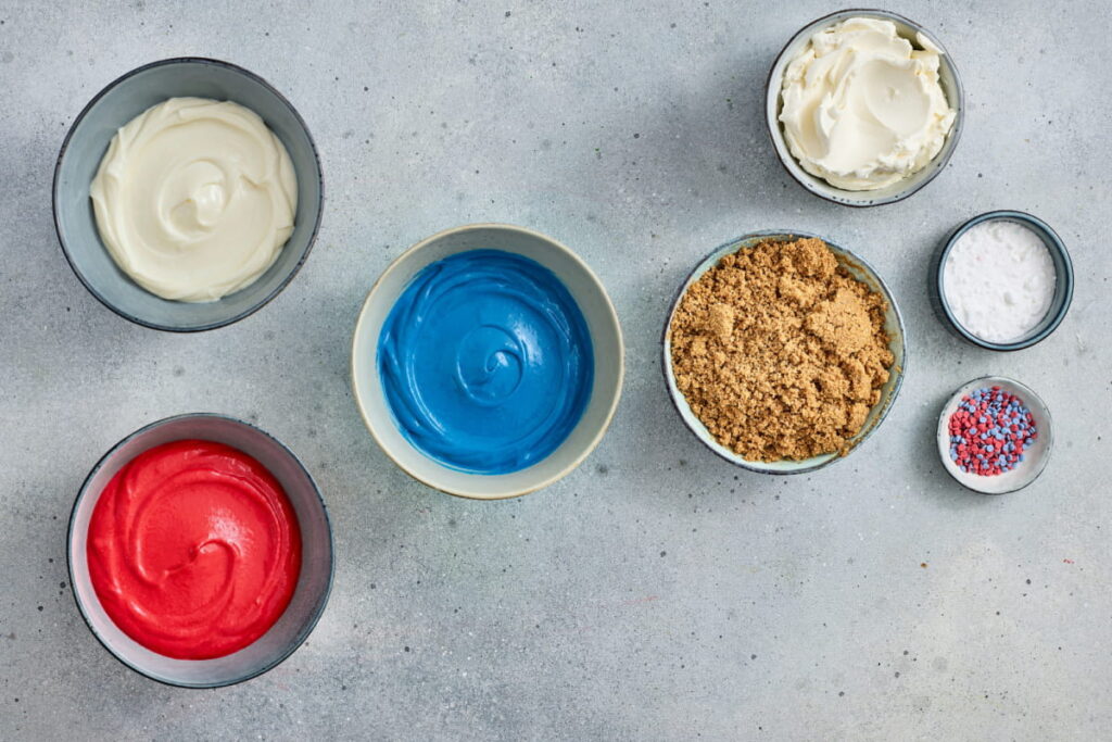Bowls of white, red, and blue frosting, brown sugar, white sugar, cream cheese, and small round sprinkles on a gray surface.