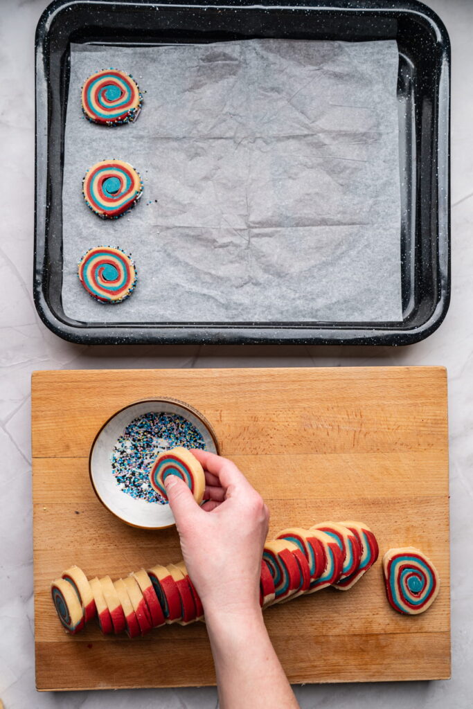 A hand dips a red, blue, and white spiral cookie into a bowl of sprinkles on a wooden board, with more cookies and a baking tray nearby.