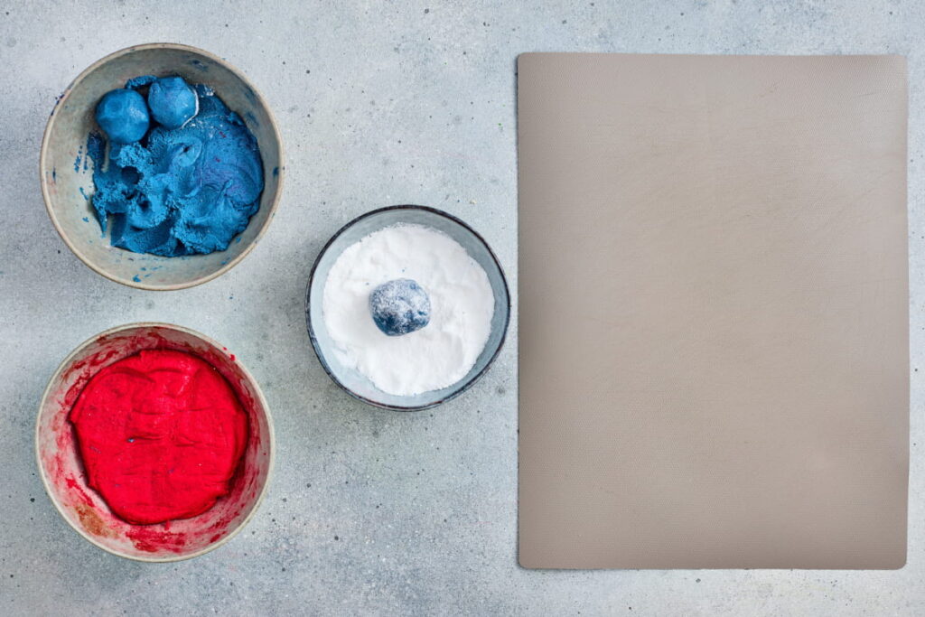 Three bowls containing blue, white, and red powder are arranged beside a closed, beige notebook on a light gray surface.