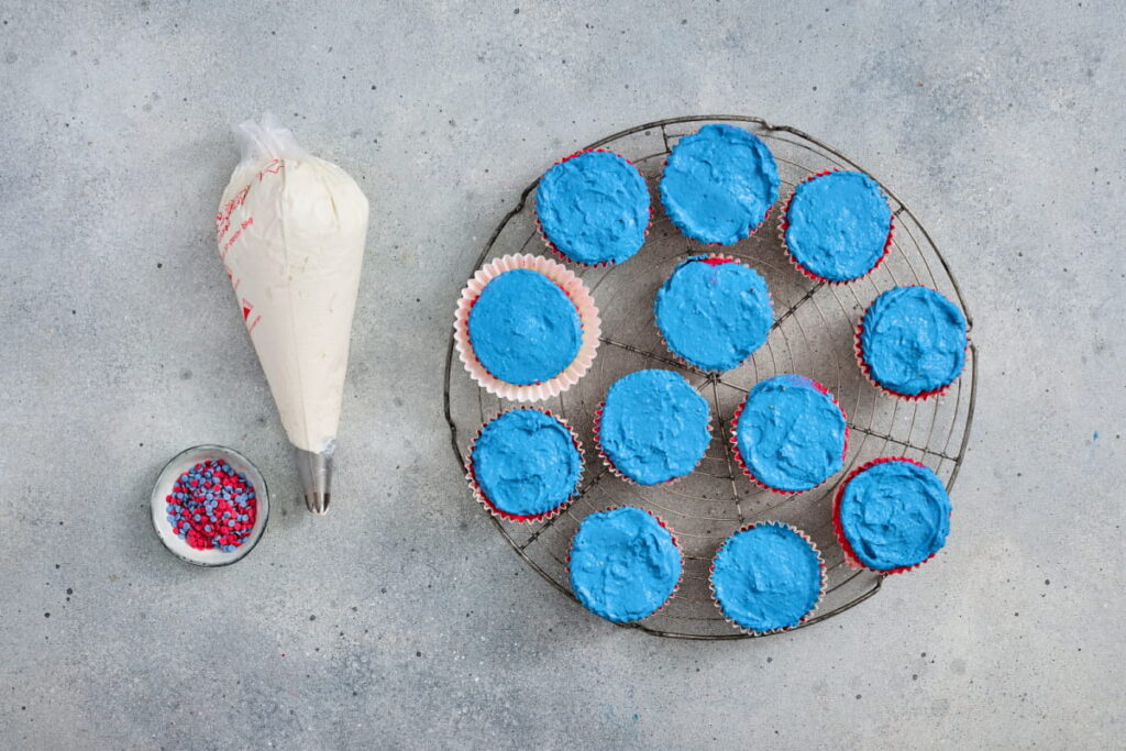 Twelve cupcakes with blue frosting on a cooling rack, one in a paper liner, with a piping bag of white frosting and a bowl of red, white, and blue sprinkles on a gray surface.