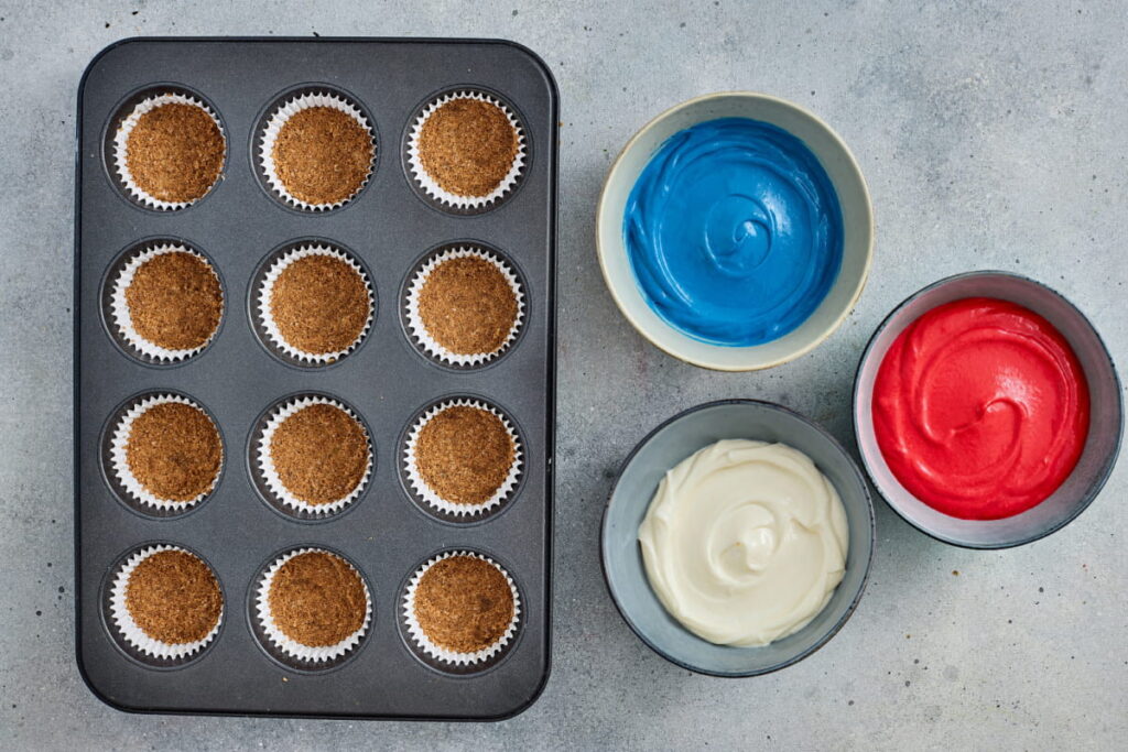 A muffin tin with 12 crust-filled cupcake liners next to bowls of blue, red, and white frosting on a light surface.