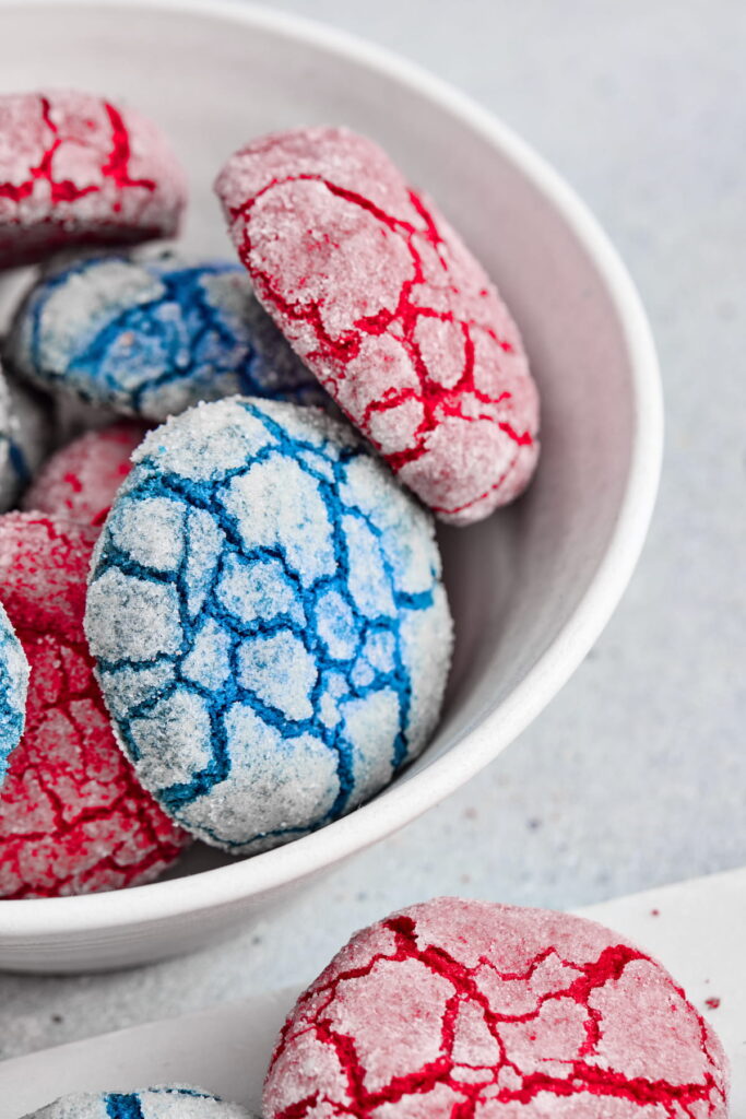 A white bowl filled with sugar-coated crinkle cookies in red and blue colors, with a cracked surface pattern.