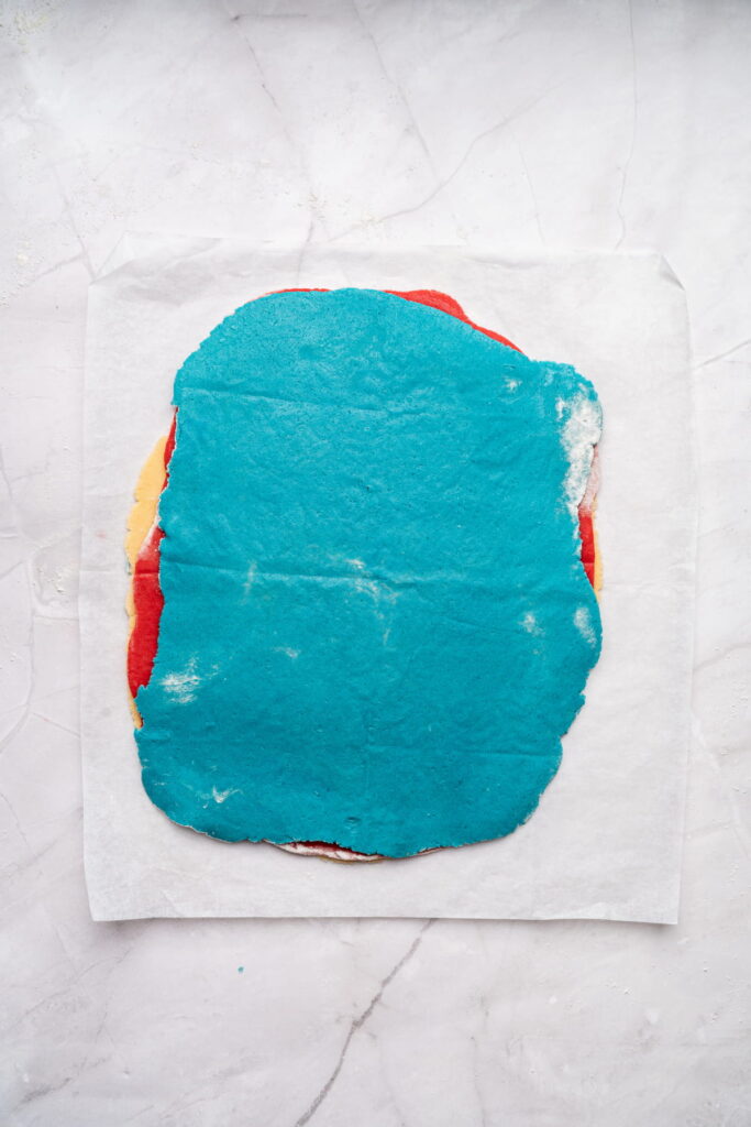 A sheet of dough layered in red, yellow, and blue colors is placed on a piece of parchment paper on a white marble surface.
