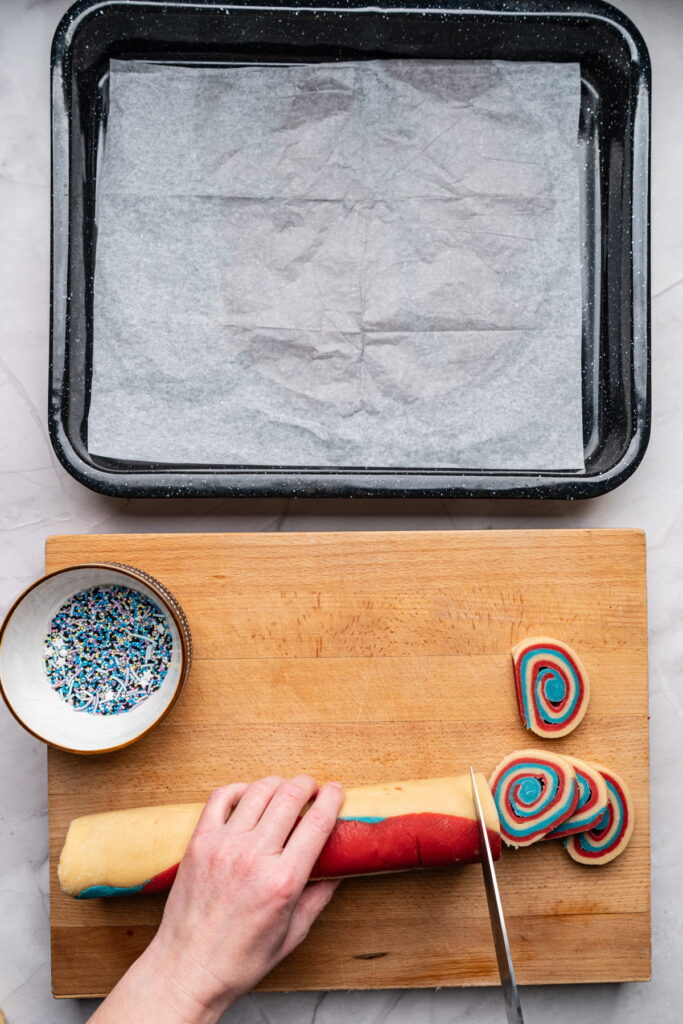 A hand slices a roll of red, blue, and white swirled cookie dough on a wooden board next to a bowl of sprinkles and a parchment-lined baking tray.