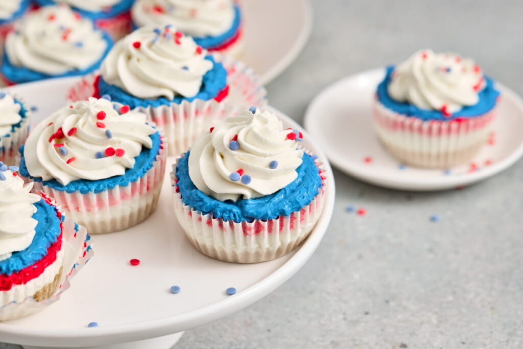 Cupcakes with white frosting, blue icing, and red, white, and blue sprinkles are displayed on a cake stand and plate.