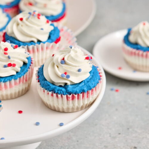 Cupcakes with white frosting, blue icing, and red, white, and blue sprinkles are displayed on a cake stand and plate.