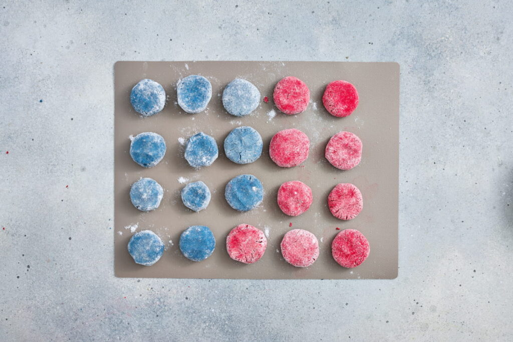 A baking sheet with four rows of round candies or cookies, half colored blue and half red, dusted with powdered sugar, on a light gray surface.
