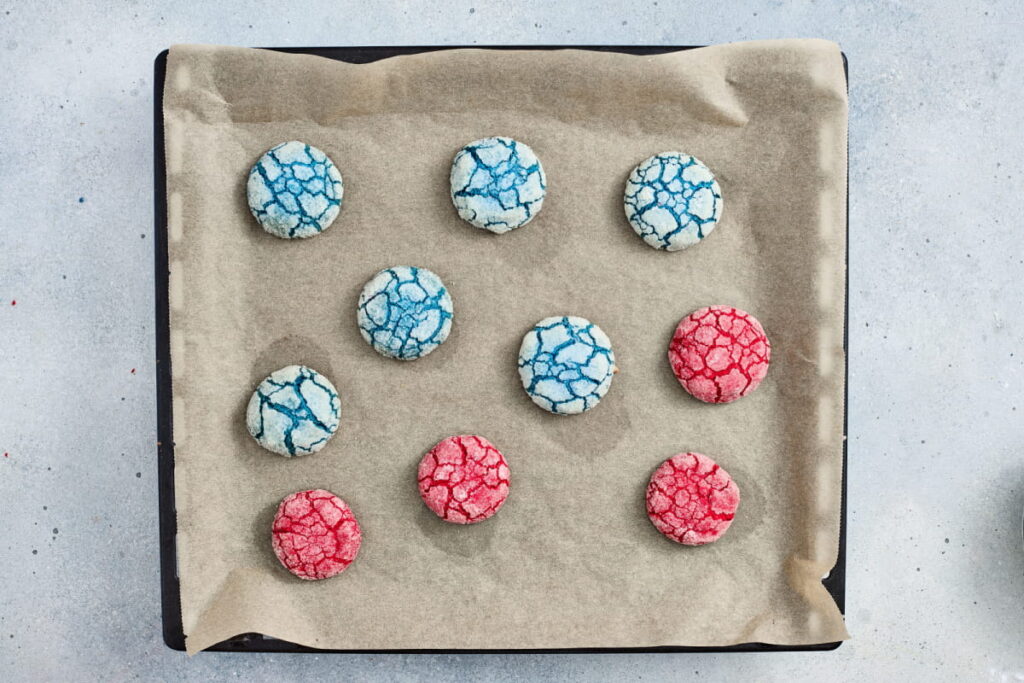 A baking tray lined with parchment paper holds eleven crinkle cookies, some blue and some red, arranged in rows on a light-colored surface.