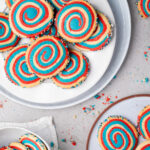 Colorful spiral cookies with red, blue, and yellow swirls are arranged on plates, with scattered crumbs and some cookies decorated with sprinkles on the edges.