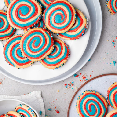 Colorful spiral cookies with red, blue, and yellow swirls are arranged on plates, with scattered crumbs and some cookies decorated with sprinkles on the edges.