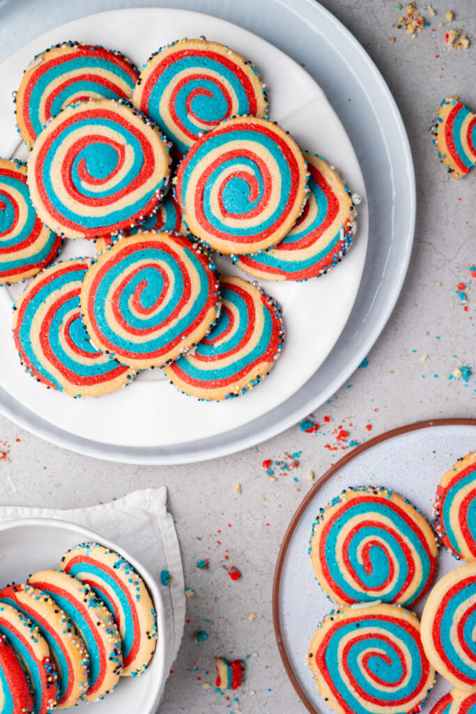 Colorful spiral cookies with red, blue, and yellow swirls are arranged on plates, with scattered crumbs and some cookies decorated with sprinkles on the edges.