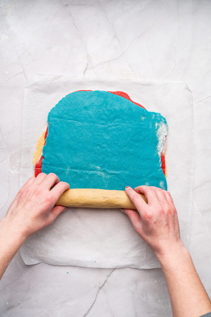 Hands rolling up multicolored layers of cookie dough, with a prominent blue layer on top, on a sheet of parchment paper.
